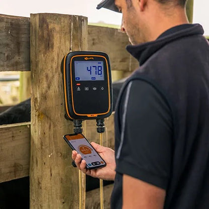 Cattle farmer in a black shirt stands beside a wooden fence post holding a smartphone, while a digital livestock scale with an orange border is mounted on the post. The scale displays a reading of 478, and a rope hangs from it.