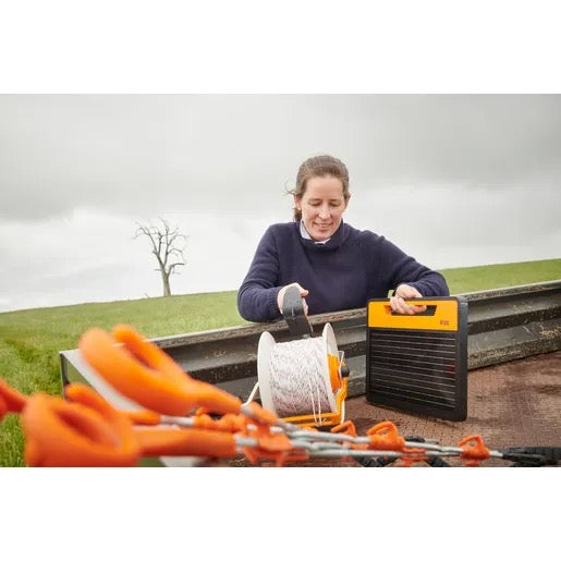 woman farmer setting up a S120 solar electric fence energizer with a spool of wire outdoors on a cloudy day on a farm