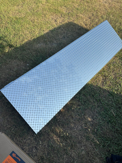 A long rectangular diamond-plate metal cattle scale platform with a textured silver finish, shown outdoors on a patch of green and brown grass.